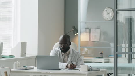 Afroamerikanischer-Arzt-Benutzt-Laptop-Und-Macht-Sich-Notizen-Bei-Der-Arbeit